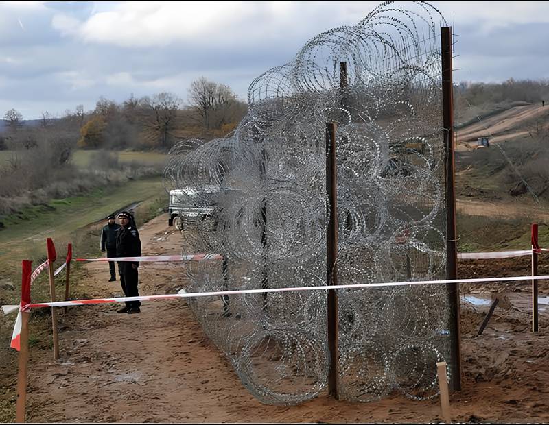 A concertina barrier in the field and two polices standing behind the barrier.